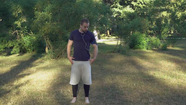 Man stretching in the park barefoot. Handsome athlete doing exercises for neck in the forest or garden. Everyday workout outdoors in the spring or summer season. Brawny young guy listening music with