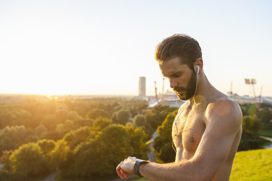 Bare chested athlete with earbuds looking on watch at sunset