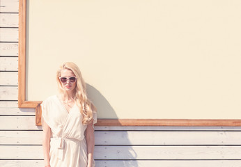 Outdoor summer sensual fashion portrait beautiful young blond woman of a white dress in sunglasses on the street on the background of a large signboard. Toned in warm colors