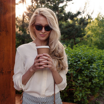 Young sexy blonde girl with long hair in sunglasses holding a cup of coffee have fun and good mood looking in camera and smiling, warm, tonning
