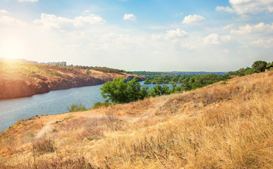 Late summer river valley, rocky hills with green trees and dry yellow grass. Blue sky with clouds, sunset.