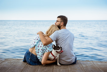 Couple sitting back to camera with French bulldog near sea.