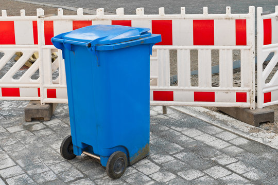 Eine blaue Mülltonne steht vor weiss-roten Absperrbaken