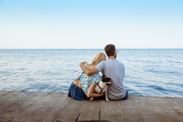 Couple sitting back to camera with French bulldog near sea.