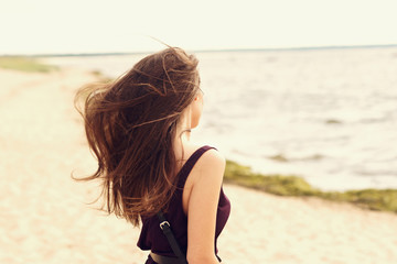 Fototapeta na wymiar Freedom - long hair brunette woman enjoying sea view at the beach. Beautiful woman in black dress standing at beach