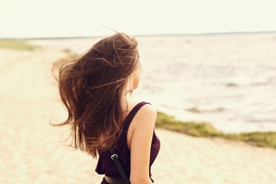 Freedom - long hair brunette woman enjoying sea view at the beach. Beautiful woman in black dress standing at beach