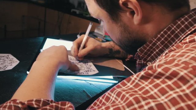 Bearded man in plaited shirt draws mandala geometrical pattern on a lightbox using copy paper