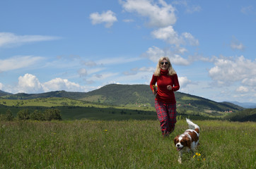 Joyful blonde woman in red with her dog (Cavalier King Charles Spaniel) on a meadow and lovely landscape in background