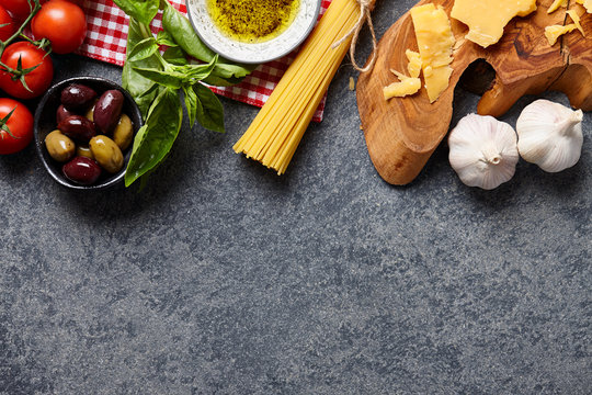 Italian food ingredients stone background with raw spaghetti, olives, basil leaves, parmesan cheese, garlic, olive oil and tomatoes.