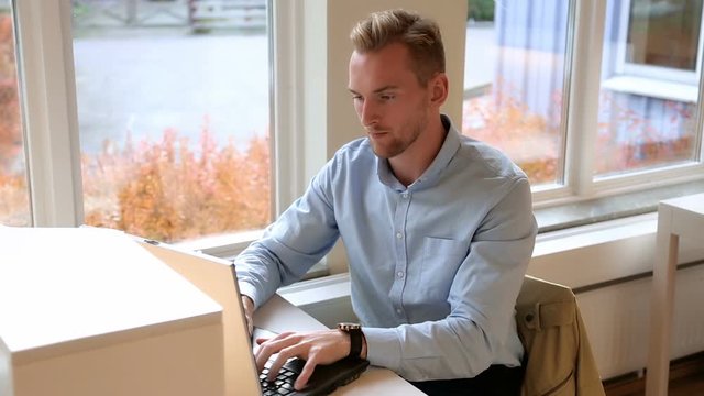 Attractive man in his 20s sitting down indoors using a laptop computer with a big window on his right with the sunlight on him. Wearing a blue shirt.