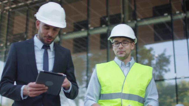 Engineer and Businessman in Hard Hat Walking, Talking, and Using Tablet Computer. Glass Building or Skyscraper under Construction on Background. Shot on RED Cinema Camera in 4K (UHD).