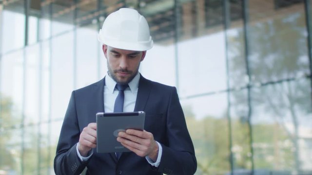 Businessman in Hard Hat Walking, Talking, and Using Tablet Computer. Glass Building or Skyscraper under Construction on Background. Shot on RED Cinema Camera in 4K (UHD).