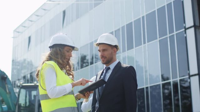 Female Engineer and Businessman in Hard Hats Having Conversation and using Tablet on Construction Site. Glass Building on Background. Shot on RED Cinema Camera in 4K (UHD).