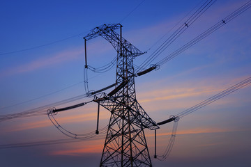 Electricity pylons, power lines and trees silhouetted against a cloudy sky at sunset.