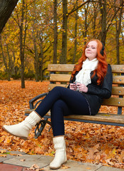 girl listen music on audio player with headphones, sit on bench in autumn city park, yellow trees...