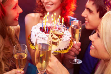 A close up of four clinking glasses against the girl holding her birthday cake among her friends