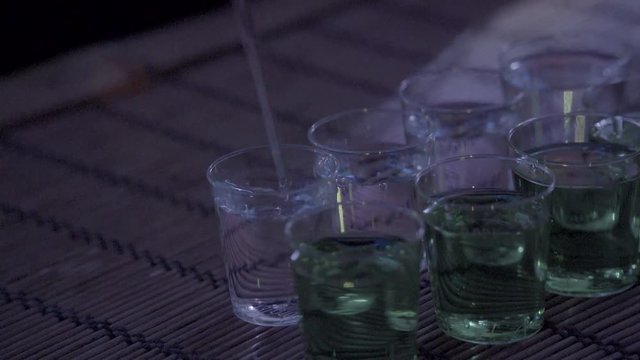 Filling a row of shot glasses with alcohol. Full glasses in the front. SLOG3, medium shot.