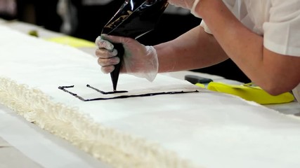 writes an inscription on a birthday cake. big cake for a birthday or a wedding, confectioner work, gloves and white uniform, qualified baker draw huge cake at the bakery, on holiday.