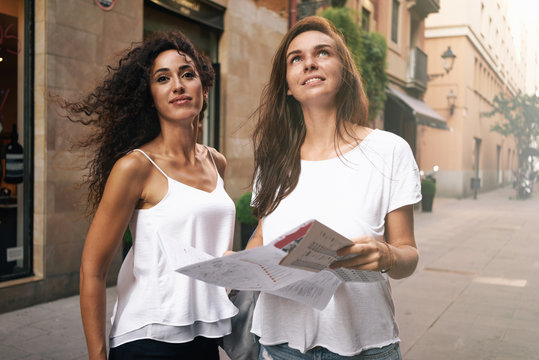 Two attractive hipster girls with long hairs are walking in the urban zone during the vacation. Friends wearing casual clothes are using map to find interesting places while travelling.