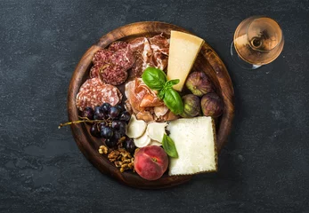  Italian antipasti snacks and glass of vine wine. Prosciutto di Parma, salami, cheese variety, figs, grapes, peach, walnuts and fresh basil on wooden serving tray over dark grunge background. Top view © sonyakamoz