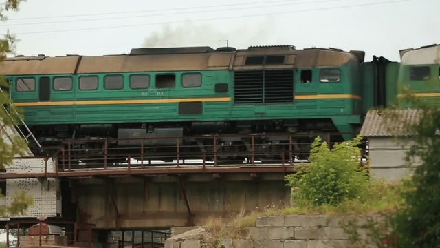 Old green train moving over metal railway bridge with grey clouded sky as background