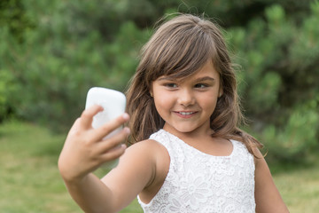 Smiling attractive little girl is doing selfie outdoors. Girl is looking at the smart phone. All potential trademarks are removed. 