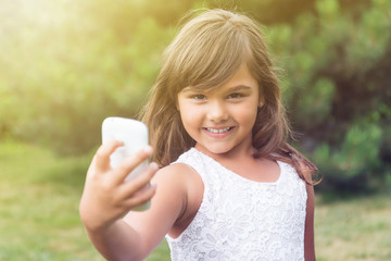 Smiling attractive little girl is doing selfie in summer light. Girl is looking at the camera holding smart phone in her hand. All potential trademarks are removed. 