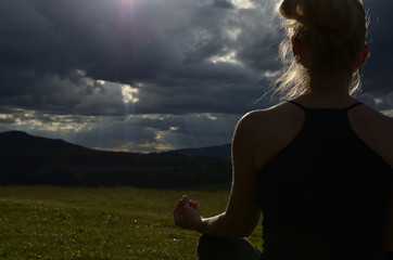 Image of a woman meditating in a field in the evening with face turned to the sun behind stormy sky