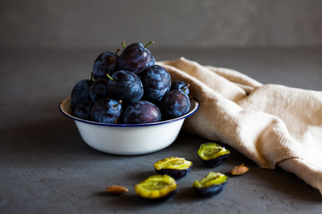 Plums in a bowl on a rural tablecloth on a gray background and half plums and plum stones on the table