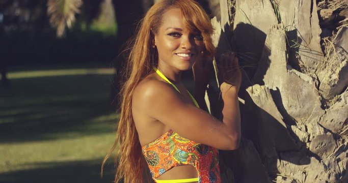 Sensual sexy young African woman standing in a colorful swimsuit holding the trunk of a tropical palm tree and smiling at the camera