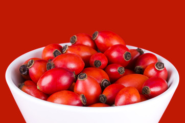Rose hips in white bowl over red, also rose haw or rose hep. Ripe red fruits of roses, used for herbal teas, jam and they can be eaten raw. One of the richest vitamin C sources available in plants.