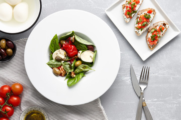 Italian salad with basil leaves, olives, capers, walnuts, mozzarella cheese and red pepper and traditional bruschetta on stone background.