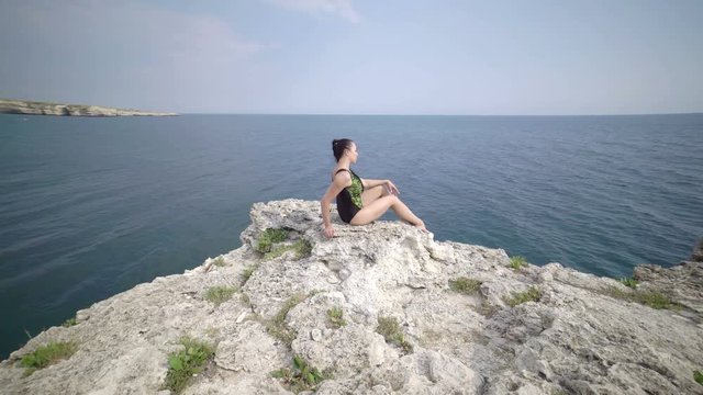 Fit woman in gymnastics leotard  posing on the edge of rock above the sea. Pole dance fitness workout outdoors. Steadicam shot 4k