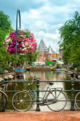 Typical Amsterdam view with bikes, canals and historical buildings. In the back, Nieuwmarkt square is dominated by the gate of the medieval city. - obrazy, fototapety, plakaty