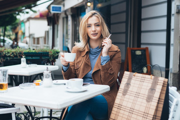 Beautiful blond woman sitting alone in cafeteria, smoking a cigarette and drinking coffee. © hedgehog94
