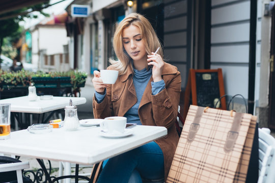 Beautiful blond woman sitting alone in cafeteria, smoking a cigarette and drinking coffee.