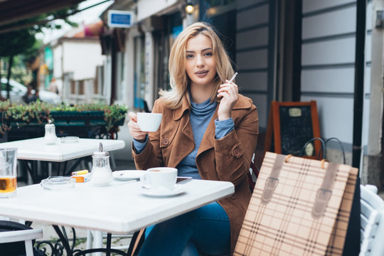 Beautiful blond woman sitting alone in cafeteria, smoking a cigarette and drinking coffee.