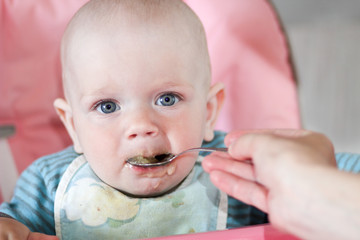 Beautiful baby eats porridge from mom's hand. He is sitting on a pink children' chair.