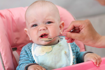 Beautiful baby eats porridge from mom's hand. He is sitting on a pink children' chair.