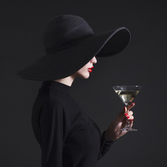 Luxury woman in a large black hat and bright lips with a glass of Martini.