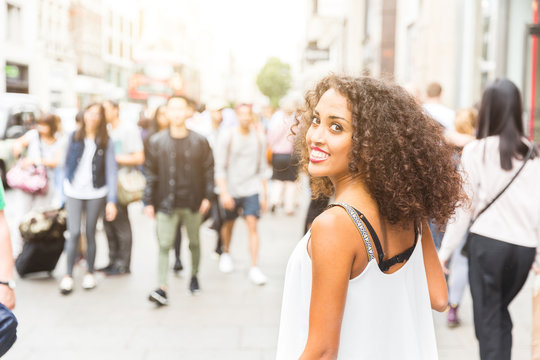 Young woman looking at camera while walking in London. Mixed race girl looking back and smiling. Blurred people on background walking on the sidewalk