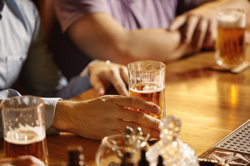 Man in shirt sitting at bar counter with his partner, celebrating anniversary of their company, drinking lager or ale. Good old friends drinking beer at pub. Selective focus on hand with glass