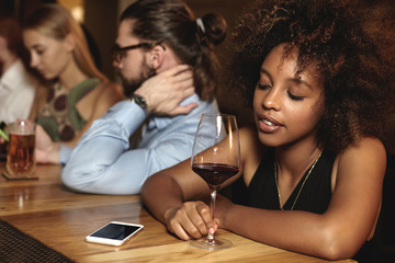 Sad pensive down hearted dark-skinned woman with glass of red wine at bar counter during New Year celebration, looking at cellphone with copy space for your advertisement, waiting for important call - Powered by Adobe