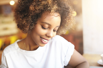 Portrait of attractive young African woman with healthy shiny skin and charming smile waiting for her friends, sitting at coffee shop. Black girl wearing white T-shirt enjoying free time at cafe