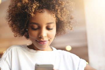 Portrait of good-looking African woman in white top browsing Internet, updating applications, using free wi-fi on electronic device, sitting at cafeteria. Black girl typing messages with mobile phone