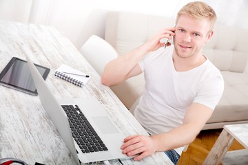 A young caucasian man working in his home office on the phone..