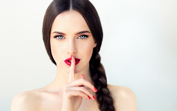 Beautiful  brunette model girl with  long braid hair . Hairstyle  pigtail  . Red lips and nails manicure .Womanl holds  finger on the  mouth surprise Shh