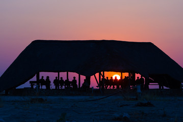 Group of tourists watching colorful sunset under shelter. Tourist resort in Africa. Backlight, silhouette, rear view, unrecognizable people.
