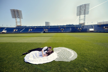 Fototapeta na wymiar Wedding couple on the football stadium is hugging each other. Beautiful girl in white dress. Man in suit. Beauty bride with groom. Female and male portrait. Woman with lace veil. Lady and guy outdoors