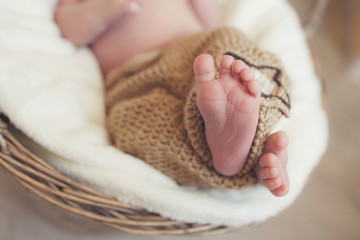 Closeup of legs of a newborn baby peeking out of light brown knitted blankets,which the baby covered from the waist up,the baby sleeps on a white soft blanket in a large wicker basket